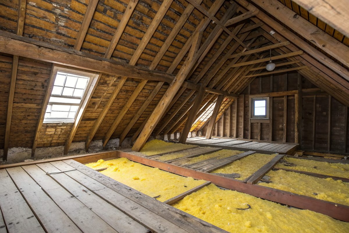 An older attic with wood rafters, plank roof boards, a small window and yellow insulation between the joists
