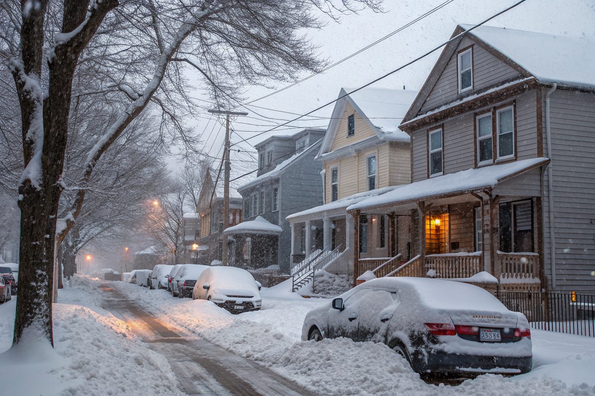 Heavy snow on the roofs, porches and parked cars along a street of older frame houses